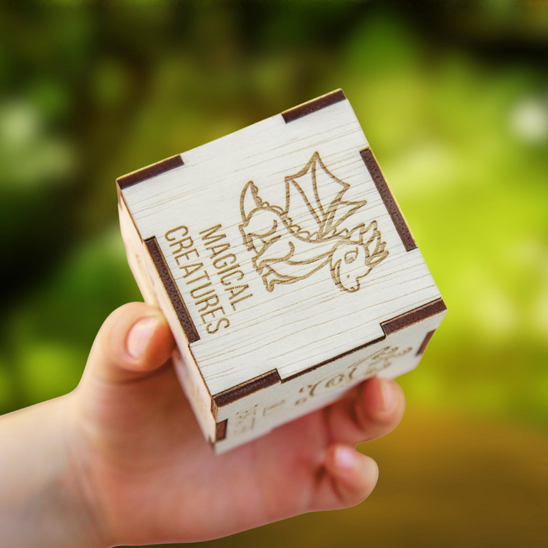 Hand holding a wooden cube with 'Magical Creatures' and dragon design against a blurred green background